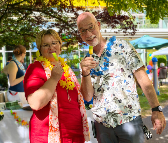 Frau im roten Kleid links und Mann im hellen Kurzarmhemd - beide haben eine Blumenkette um den Hals und ein Eis am Stil in der Hand.
