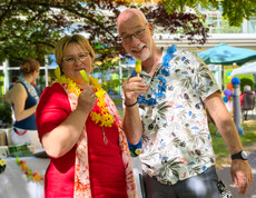 Frau im roten Kleid links und Mann im hellen Kurzarmhemd - beide haben eine Blumenkette um den Hals und ein Eis am Stil in der Hand.
