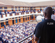 Ein Mann im FLEM-T-Shirt steht auf einer oberen Etage und spricht durch ein Megafon zu einer großen Gruppe von Schüler*innen, die dicht gedrängt im Innenhof einer mehrstöckigen Schule stehen und zuhören.