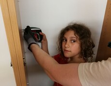 Ein Kind unterstützt beim Aufbau eines Schranks und verwendet unter Anleitung einen Akkuschrauber bei handwerklichen Arbeiten.