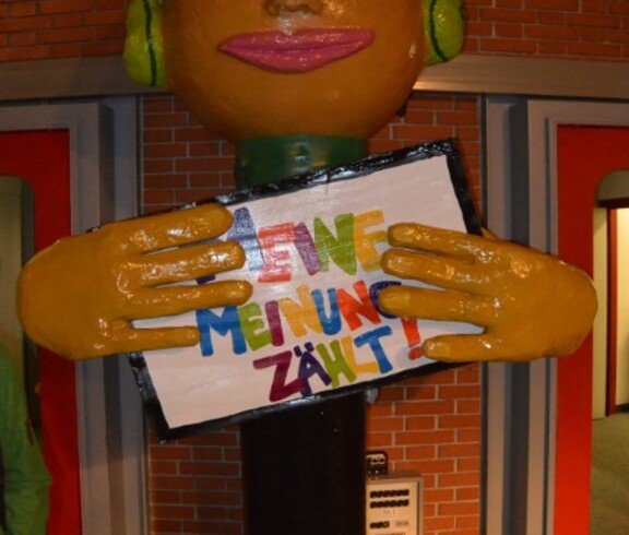 Eine große Figur hält eine Botschaft mit der Aufschrift Meine meine Meinung zählt. Die Figur stellt einen Jungen dar und trägt grüne Kopfhörer.