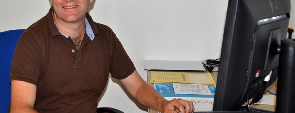 Ein Herr mittleren Alters sitzt an einem Schreibtisch vor einem Computer. Er trägt ein braunes Polo Shirt und hält seine rechte Hand auf der Computermaus. 