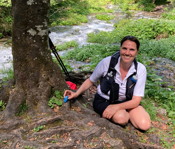 Frau legt bemalten Stein bei einem Baum neben einem Fluss ab.