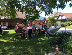 Bewohner*innen sitzen beim Grillfest im Garten des St. Hemma Hauses unter großen Bäumen an Festbänken.