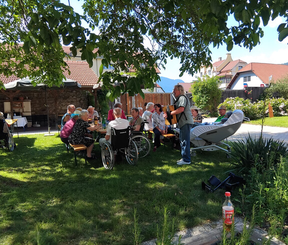 Bewohner*innen sitzen beim Grillfest im Garten des St. Hemma Hauses unter großen Bäumen an Festbänken.