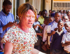 Eine Frau in einem geblümten Kleid spricht gestikulierend zu einer großen Gruppe von Schüler*innen, die eng beisammenstehen und aufmerksam zuhören; im Hintergrund stehen weitere Erwachsene und Jugendliche im Schulflur.