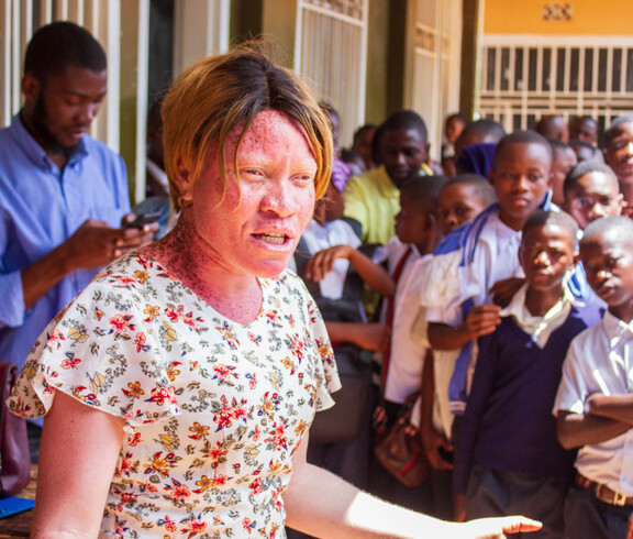 Eine Frau in einem geblümten Kleid spricht gestikulierend zu einer großen Gruppe von Schüler*innen, die eng beisammenstehen und aufmerksam zuhören; im Hintergrund stehen weitere Erwachsene und Jugendliche im Schulflur.