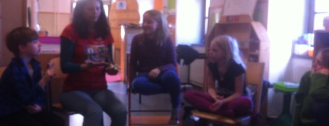 Kinder sitzen mit einer Betreuungsperson im Stuhlkreis und nehmen gemeinsam an einer Lern- und Gesprächsrunde im Klassenraum teil.