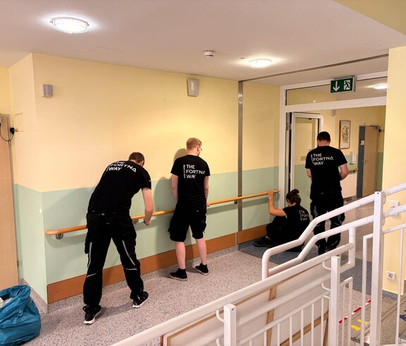 Vier Männer – mit dem Rücken zur Kamera – tragen Firmen-Shirts mit der Aufschrift „The Fortna Way“. Sie montieren einen langen Haltegriff entlang der frisch ausgemalten Wand.