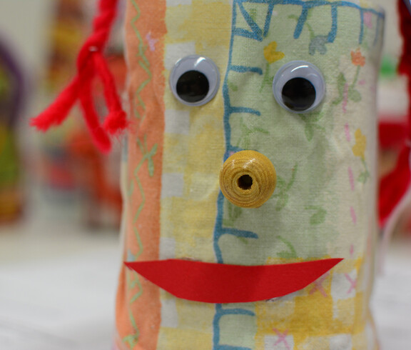 Eine Figur, die aus einem Glas gemacht ist, hat zwei Kulleraugen, einen roten Mund aus Papier und eine gelbe, schneckenartige Nase.
