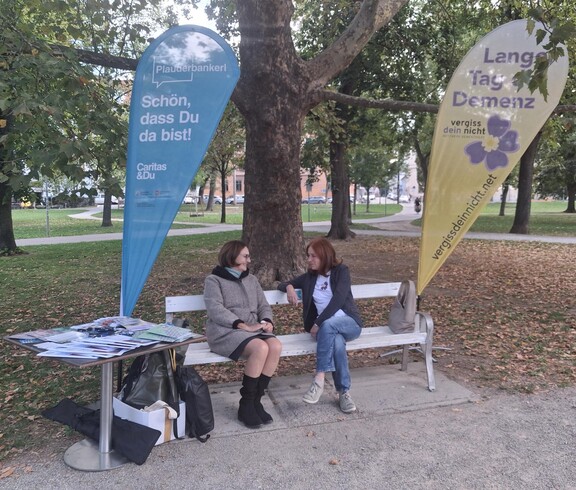 An einem Informationsstand zum Tag der Demenz sitzen zwei Personen auf einer Bank neben Broschüren und Informationsmaterialien, eingerahmt von zwei Fahnen in einer Parkanlage.