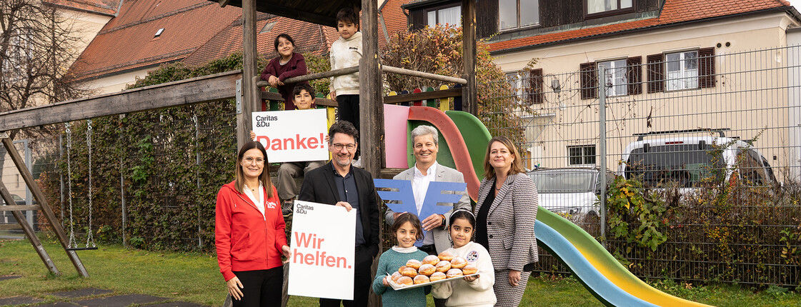 Vertreter*innen der Volksbank mit Caritas-Team und Kindern vor Rutsche im Garten eines Lerncafés, halten Schilder mit „Wir helfen“ und „Danke!“