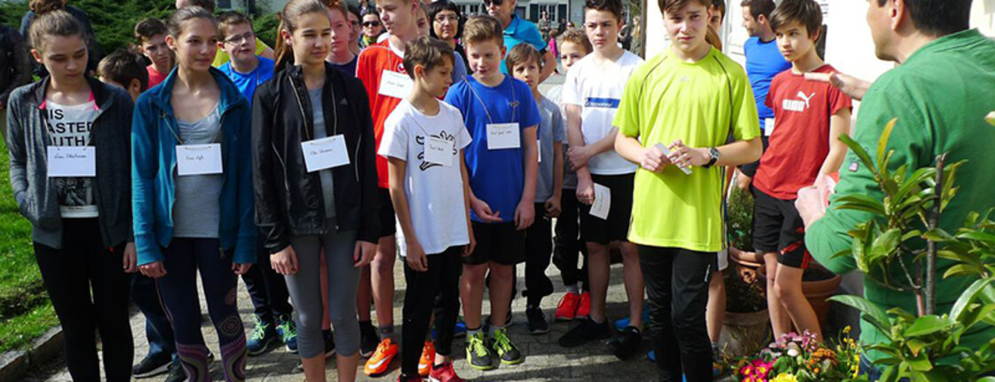 Mehrere Menschen stehen hintereinander aufgestellt. Alle tragen Sportkleidung und schauen in die Richtung eines Mannes, der etwas erklärt.