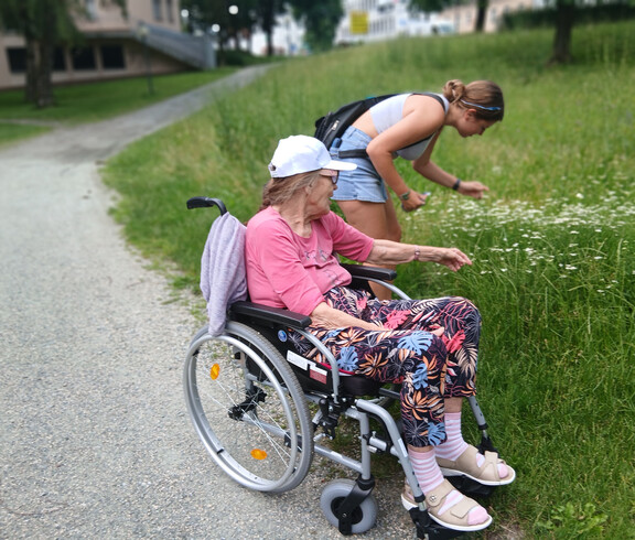 Bewohnerin im Rollstuhl und Schülerin betrachten gemeinsam Blumen am Wegesrand im Park.