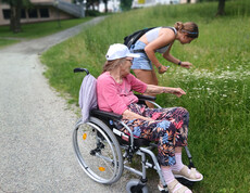 Bewohnerin im Rollstuhl und Schülerin betrachten gemeinsam Blumen am Wegesrand im Park.