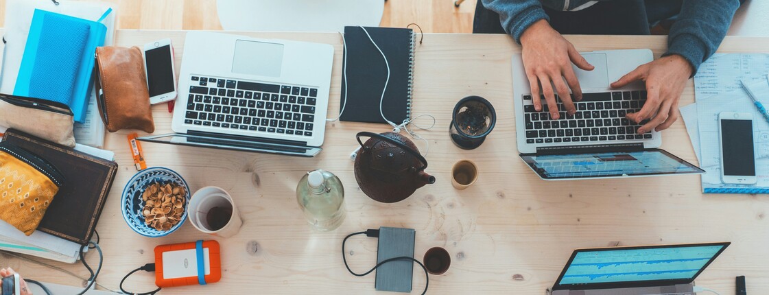 Obersicht eines modernen Schreibtisches aus hellem Holz, an dem fünf Personen mit Laptops und weiteren Bürogeräten arbeiten.