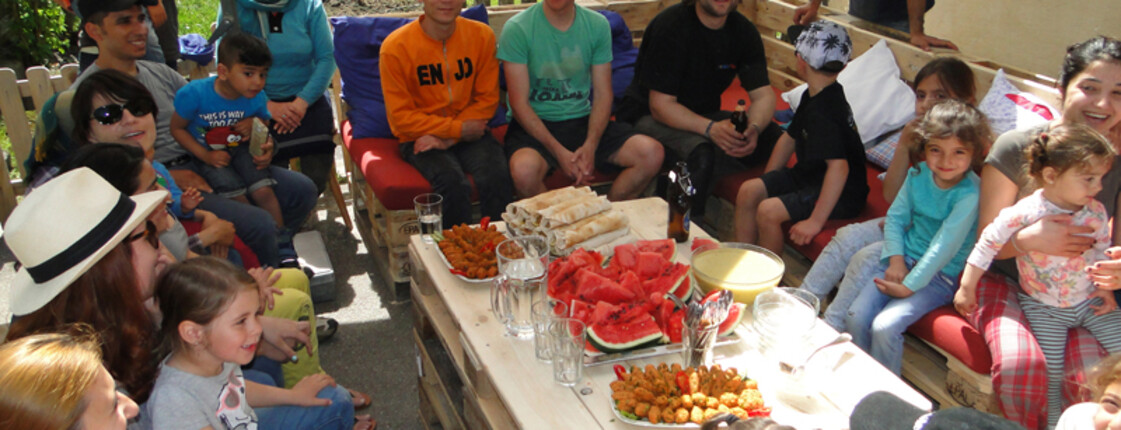 Viele Menschen sitzen im Freien auf selbstgemachten Holzpaletten Sofas. in der Mitte befindet sich ein Holztisch mit verschiedenen Häppchen.