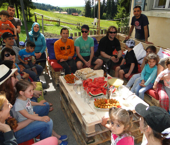 Viele Menschen sitzen im Freien auf selbstgemachten Holzpaletten Sofas. in der Mitte befindet sich ein Holztisch mit verschiedenen Häppchen.
