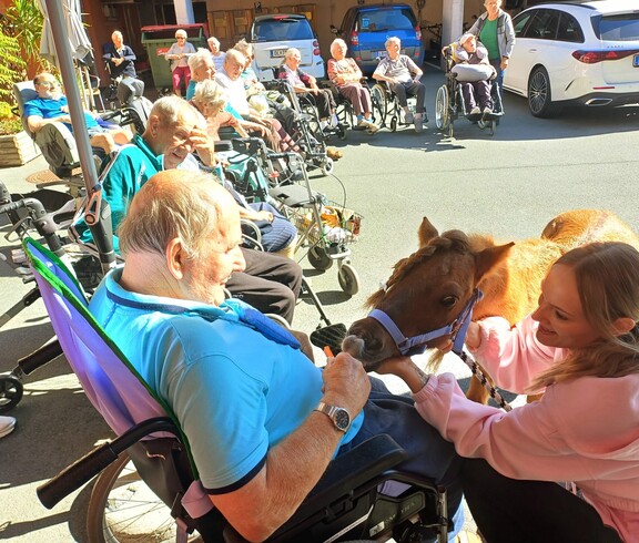 Ein Bewohner im Rollstuhl füttert Pony Maxi aus der Hand, während weitere Bewohnerinnen und Bewohner die Begegnung im sonnigen Innenhof des Pflegewohnhauses beobachten.