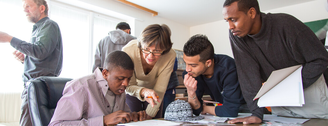 Flüchtlinge aus Afrika und dem nahen Osten lernen Deutsch. Sie sitzen gespannt und konzentriert an einem Tisch und blicken auf ihre Unterlagen. Eine Lehrerin hilft ihnen dabei den Stoff besser aufzunehmen und steht für jede Frage offen. 