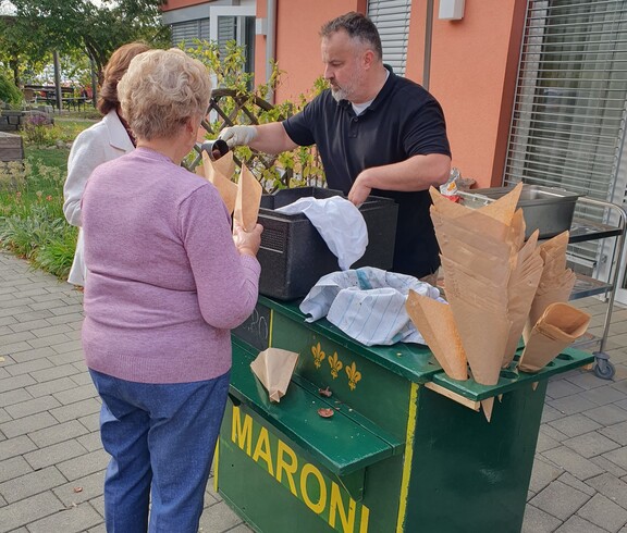 An einem Maronistand werden frisch gebratene Maroni in Papiertüten ausgegeben, während mehrere Personen im Freien auf der Terrasse zusammenkommen.