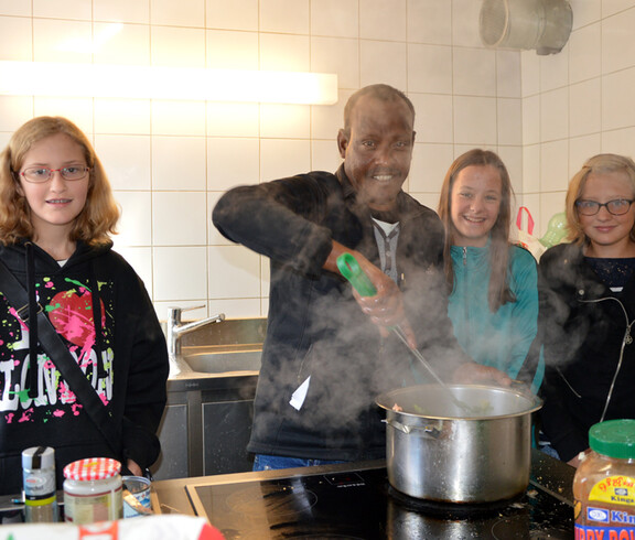 Drei Kinder und ein Herr sind in einer Küche. Der Herr kocht etwas.