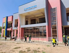 Das neue zweistöckige Schulgebäude mit farbiger Fassade, vor dem Schulkinder in Uniform entlanggehen.