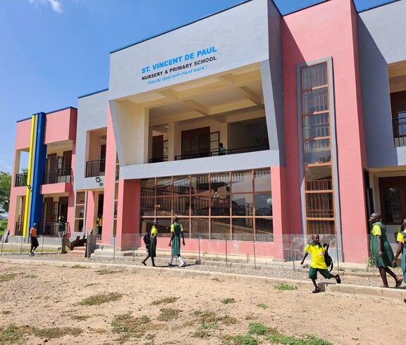 Das neue zweistöckige Schulgebäude mit farbiger Fassade, vor dem Schulkinder in Uniform entlanggehen.