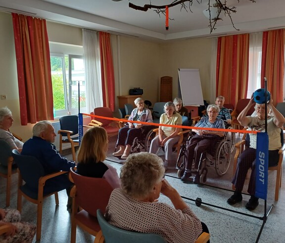 Einige Bewohner*innen des Caritas Pflegewohnhauses Lannach sitzen auf Stühlen oder in Rollstühlen und spielen gemeinsam über ein niedriges Netz mit einem Softball. Die Szene zeigt Freude, Bewegung und gemeinsames Erleben.