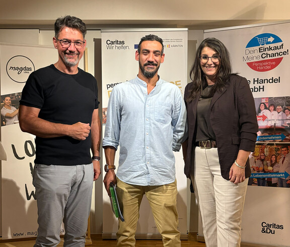 Ein Teilnehmer steht mit Caritasdirektor und Projektleiterin bei einer Zertifikatsverleihung vor Roll-ups von Caritas und Perspektive Handel.