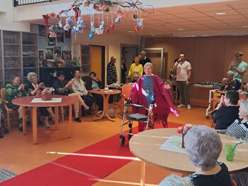 Eine Person präsentiert mit einer Gehhilfe auf einem roten Laufsteg eine Modeschau im barrierefreien Gemeinschaftsraum des Pflegewohnhauses, während das Publikum ringsum an Tischen sitzt.