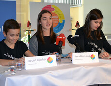 Pressekonferenz der Jugendbotschafter. Drei junge Menschen sitzen an einem Tisch mit Mikrofonen und geben einen Vortrag.