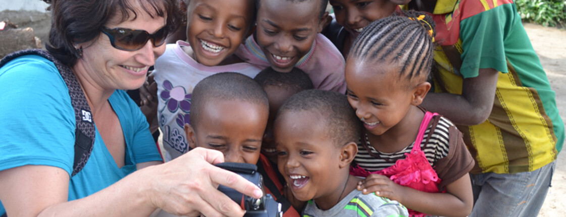 Caritas-Mitarbeiterin Elke Kager zeigt äthiopischen Kindern Fotos auf ihrer Kamera. Die Kinder schauen gespannt und sind erfreut. Elke trägt eine blaue Bluse und die Kinder sind bunt, in unterschiedlichen Farben gekleidet. 