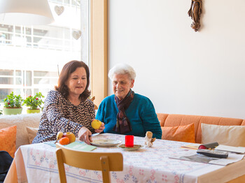 Eine Frau mittleren Alters sitzt mit einer alten Dame am Tisch. Die jüngere Dame hält einen Apfel in der Hand, den sie aufschneiden will.