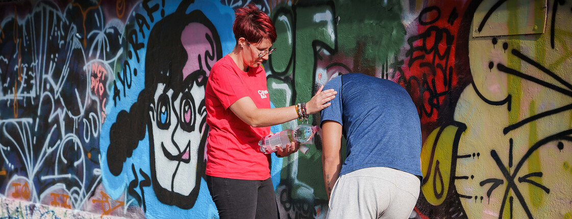 Hilfsperson reicht einer erschöpften Person an einer Graffitiwand Wasser bei großer Hitze und stützt sie im Schatten.