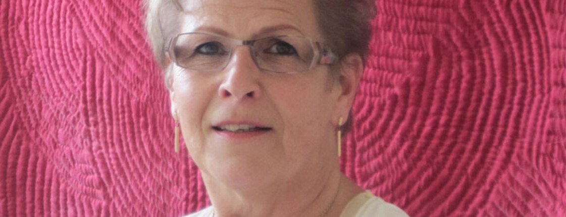 Eine Dame steht für ein Portrait vor einem pinken Hintergrund. Sie trägt eine Brille und goldenen Schmuck.