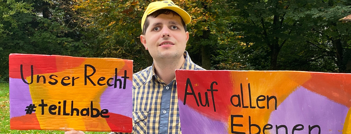 Ein Mann mit gelber Kappe hält zwei Schilder in der Hand. 