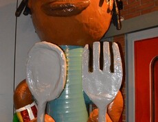 Eine Figur, die einem Menschen ähnelt, hält Löffel und Gabel in Händen. Sie ist überdemensioniert und hat dunkle Hautfarbe und hat eine Kopfbedeckung. 