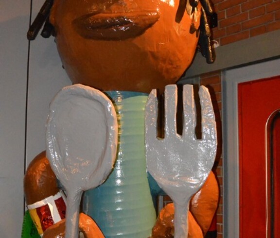 Eine Figur, die einem Menschen ähnelt, hält Löffel und Gabel in Händen. Sie ist überdemensioniert und hat dunkle Hautfarbe und hat eine Kopfbedeckung. 