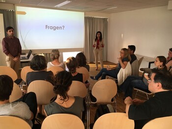 Mehrere Personen verfolgen einen Vortrag in einem Seminarraum. Zwei Personen stehen vor einer Präsentationsfolie und beantworten Fragen aus dem Publikum. 