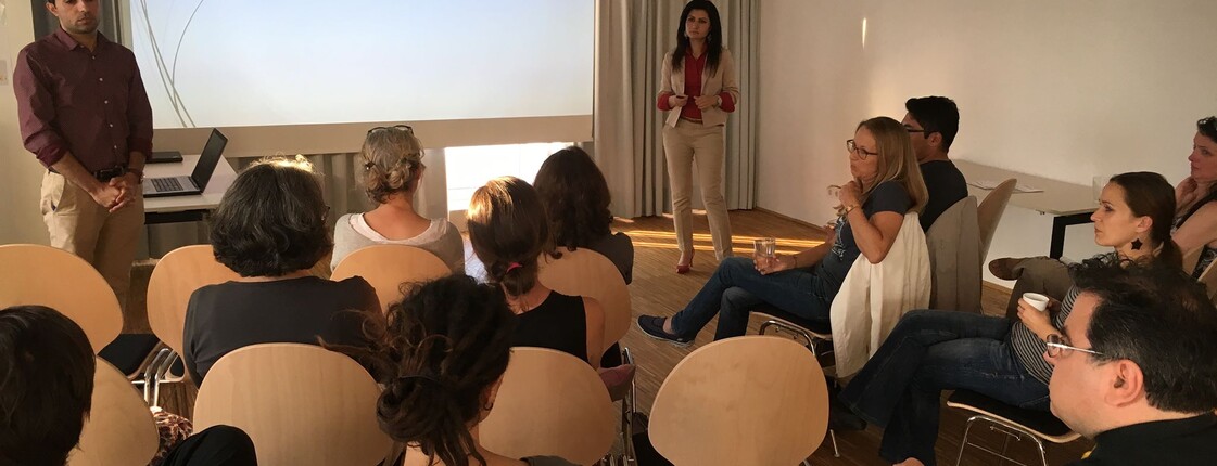 Mehrere Personen verfolgen einen Vortrag in einem Seminarraum. Zwei Personen stehen vor einer Präsentationsfolie und beantworten Fragen aus dem Publikum. 