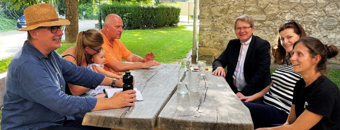 Menschen sitzen an einem Holztisch im schattigen Garten neben der Pfarrkirche und kommen zur Eröffnung der Schattenoase zusammen.