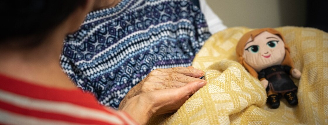 Eine Person hält im Hospiz die Hand eines bettlägerigen Menschen; auf der gelben Decke liegt eine kleine Stoffpuppe.