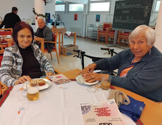 Zwei ältere Personen sitzen an einem Tisch im Speisesaal und genießen Essen und Getränke. Im Hintergrund ist eine Tafel mit der Aufschrift "Willkommen zum Weihnachtsbasar" zu sehen.