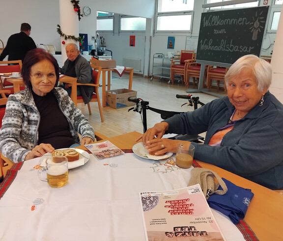 Zwei ältere Personen sitzen an einem Tisch im Speisesaal und genießen Essen und Getränke. Im Hintergrund ist eine Tafel mit der Aufschrift "Willkommen zum Weihnachtsbasar" zu sehen.