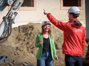 Zwei Personen mit Schutzhelmen auf einer Baustelle für Caritas-Pflegeeinrichtungen, daneben ein Bagger.