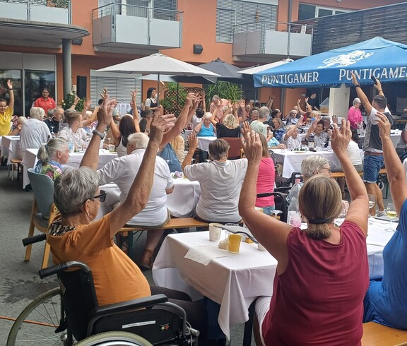 Beim Sommerfest im Innenhof des Pflegewohnhauses sitzen zahlreiche Bewohnerinnen und Bewohner an festlich gedeckten Tischen und heben gemeinsam die Arme, während das Musikprogramm für Unterhaltung sorgt.