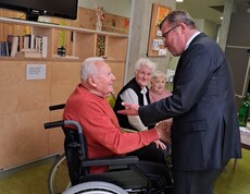 Ein Bewohner im Rollstuhl begrüßt Weihbischof Johannes Freitag mit einem Handschlag, während zwei weitere Bewohnerinnen bzw. Bewohner die Begegnung im Aufenthaltsbereich des Caritas Pflegewohnhauses verfolgen.