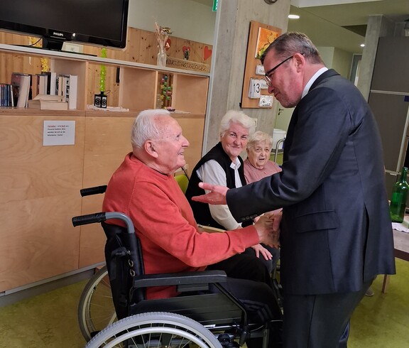 Ein Bewohner im Rollstuhl begrüßt Weihbischof Johannes Freitag mit einem Handschlag, während zwei weitere Bewohnerinnen bzw. Bewohner die Begegnung im Aufenthaltsbereich des Caritas Pflegewohnhauses verfolgen.