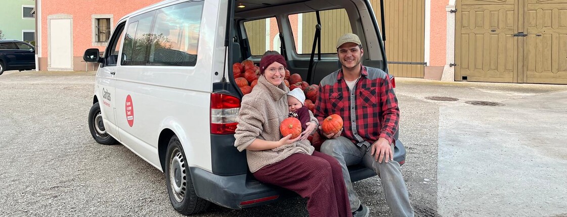 Frau und ein Mann sitzen mit Banby im offenen Kofferaum von einem Kleinbus und halten Kürbisse in der Hand
